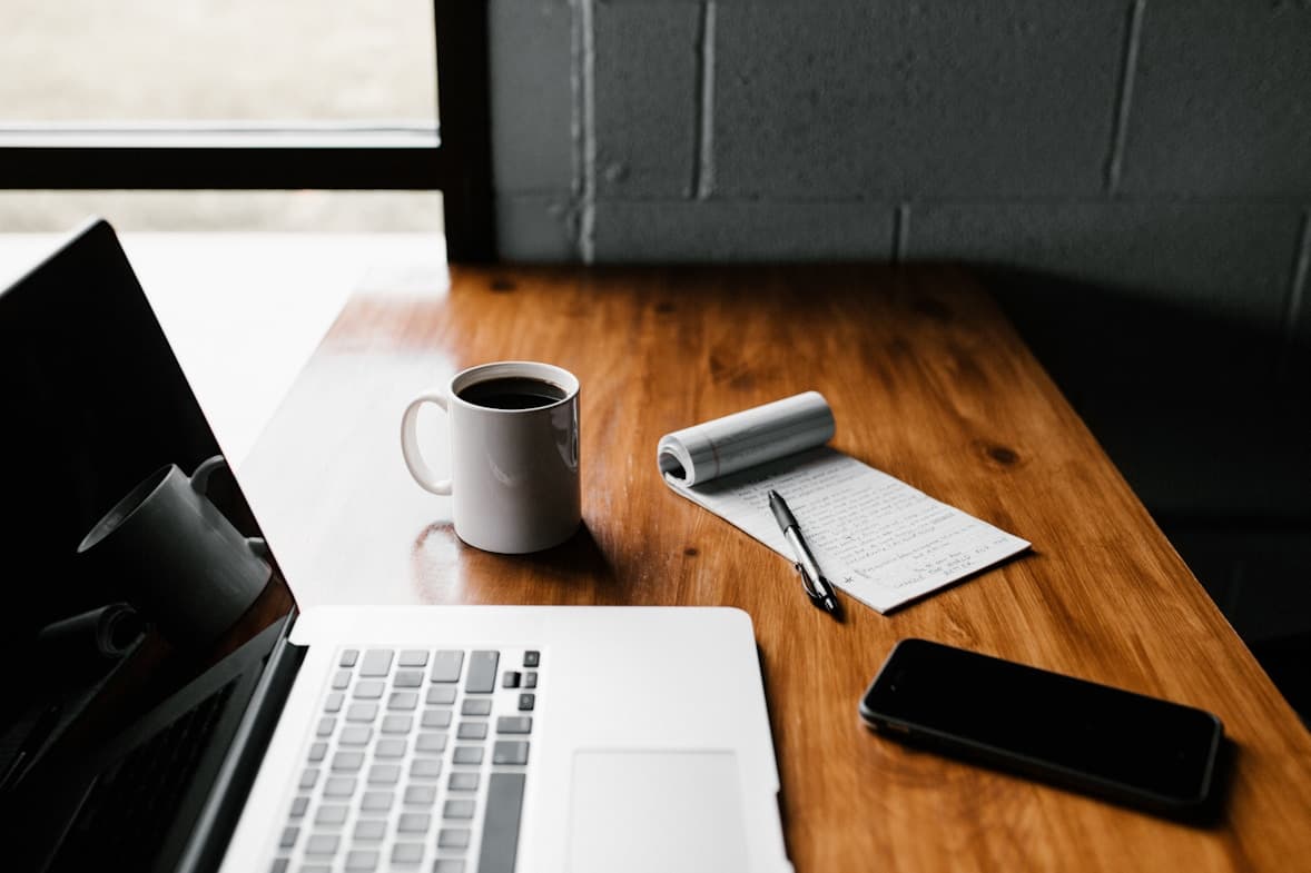 A laptop, notepad, phone and mug of coffee on a wooden desk